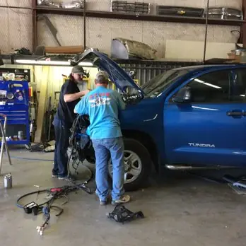 Two technicians working under the hood of a blue Toyota Tundra inside the Acme Body Shop garage
